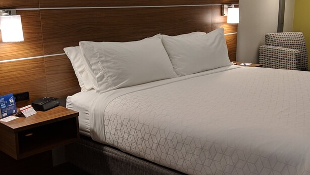 A hotel room with a wooden headboard, a bed with white bedding, a nightstand with a phone and a sign, and a chair. - Holiday Inn Express & Suites Claypool Hill (Richlands Area) by IHG (Pounding Mill)