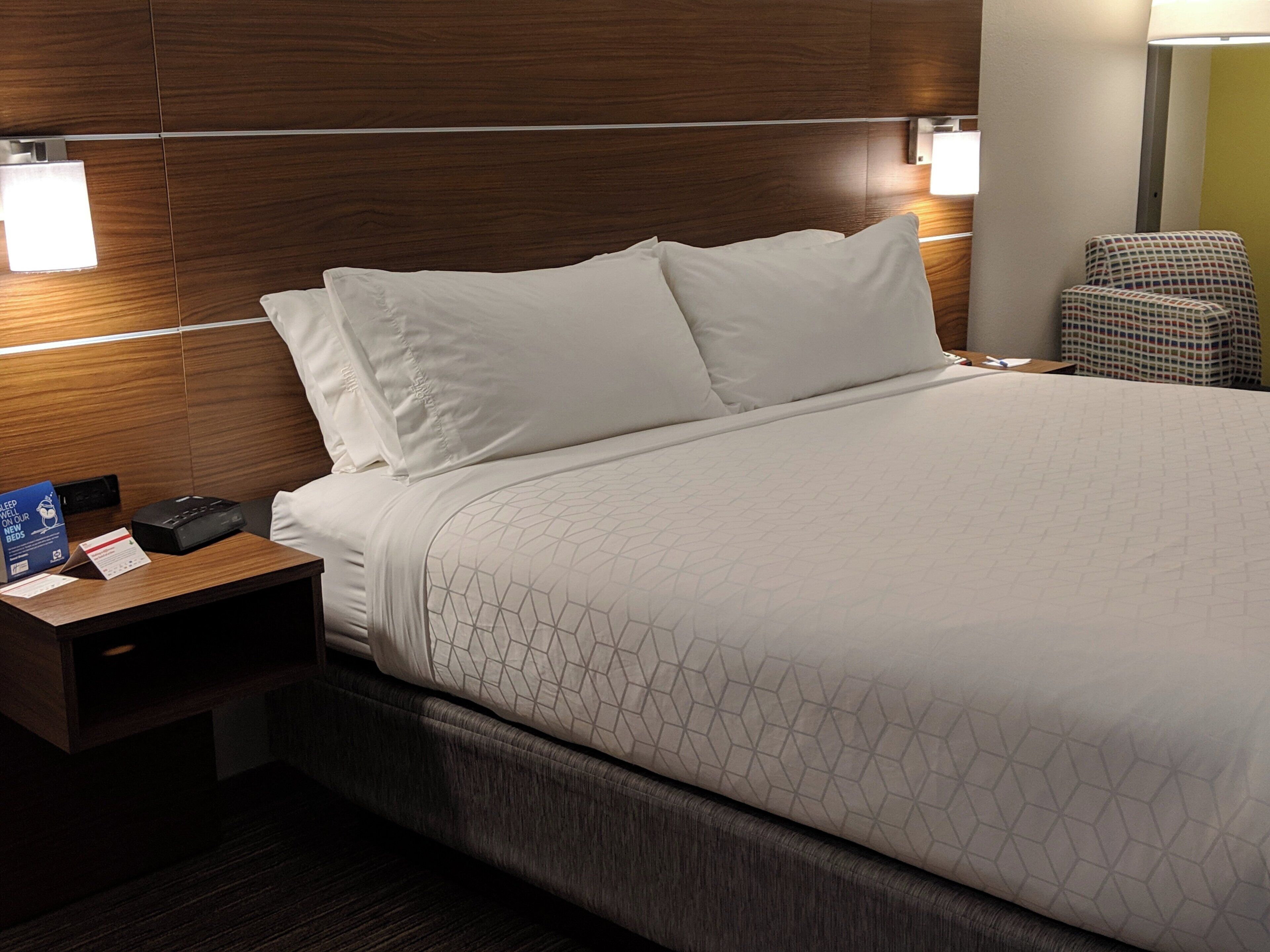 Una habitación de hotel con cabecera de madera, cama con ropa blanca, mesita de noche con teléfono y un cartel, y una silla.