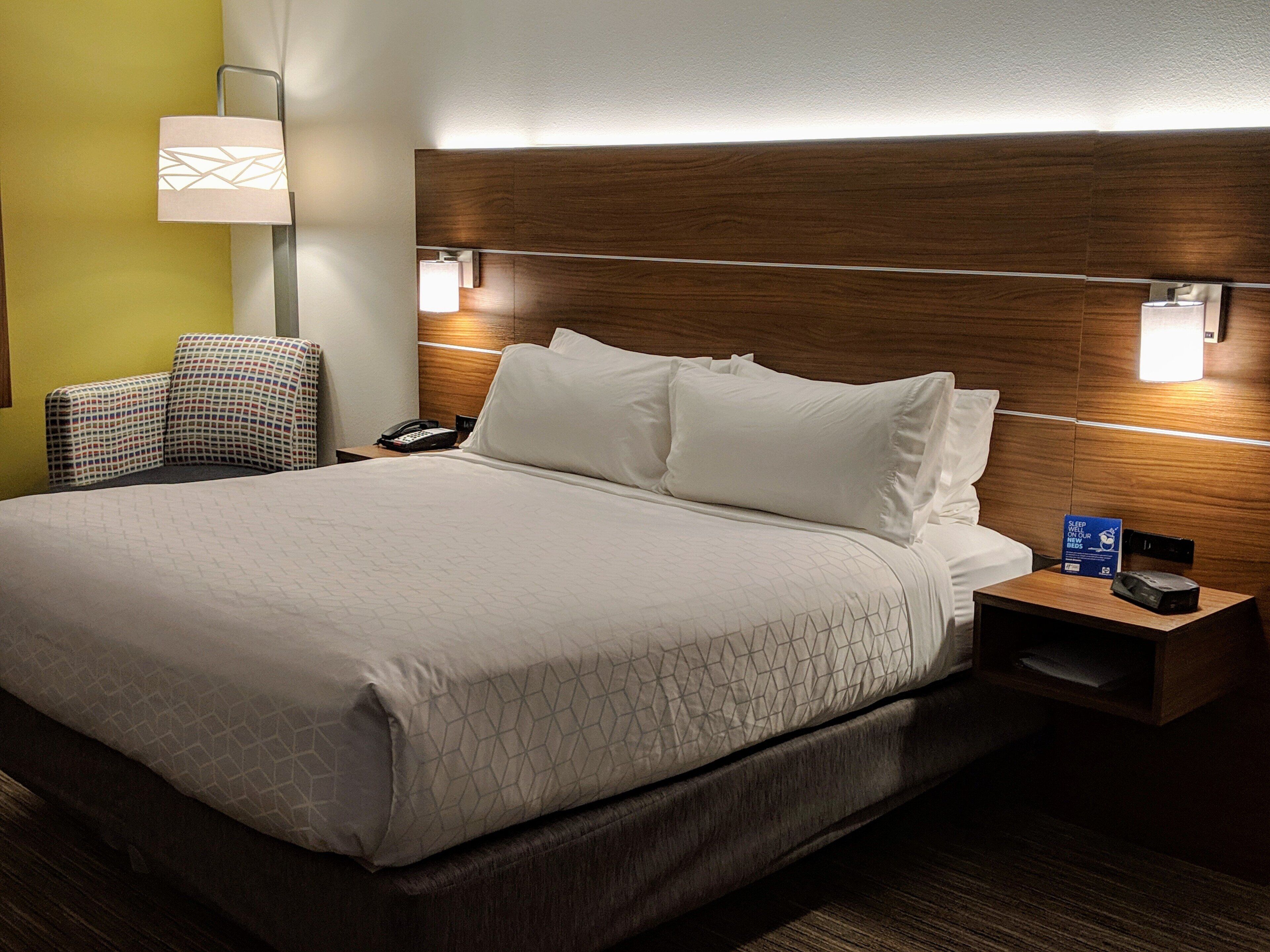 Habitación de hotel con una cama grande, una mesita de noche con lámpara, una silla y un cartel sobre camas nuevas.