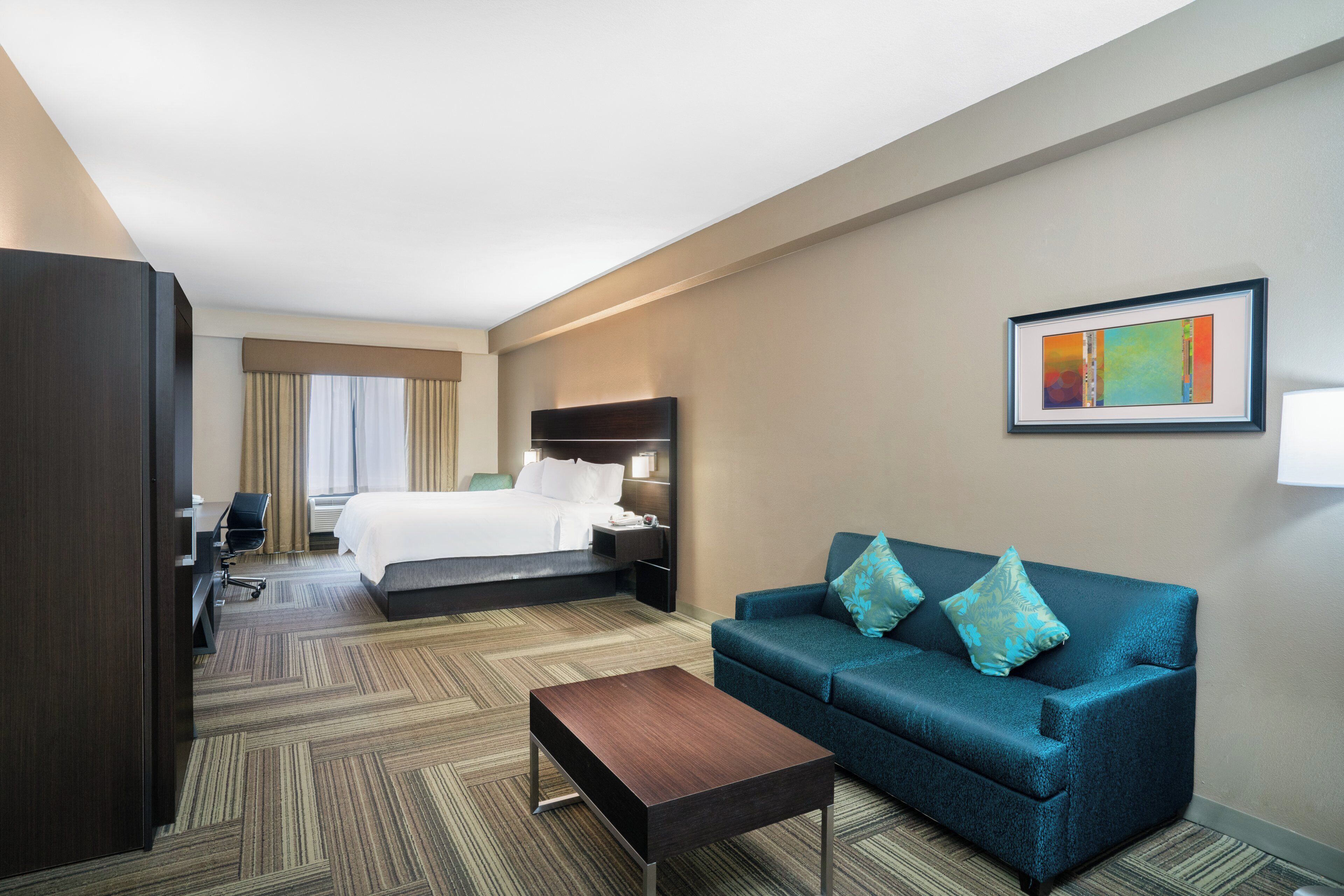 A hotel room with a bed, a sofa, a coffee table, a desk with a chair, and a painting on the wall.