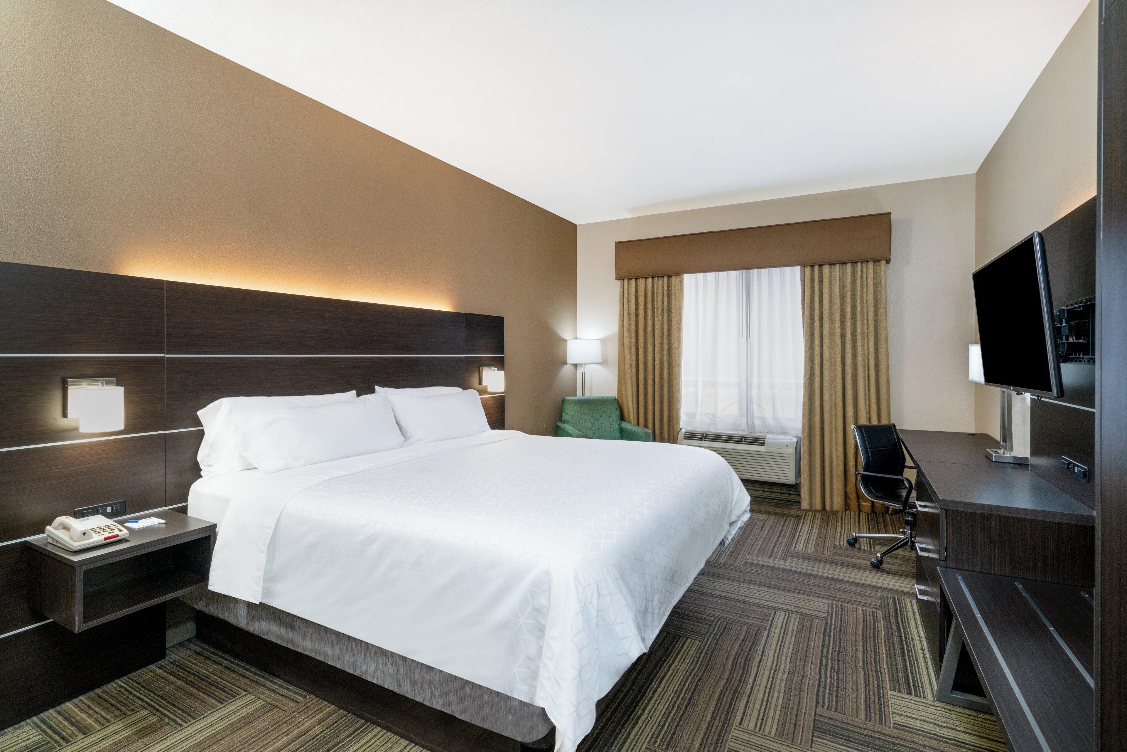 Habitación de hotel con una cama grande, un escritorio con televisor, una silla, una lámpara y una ventana con cortinas.