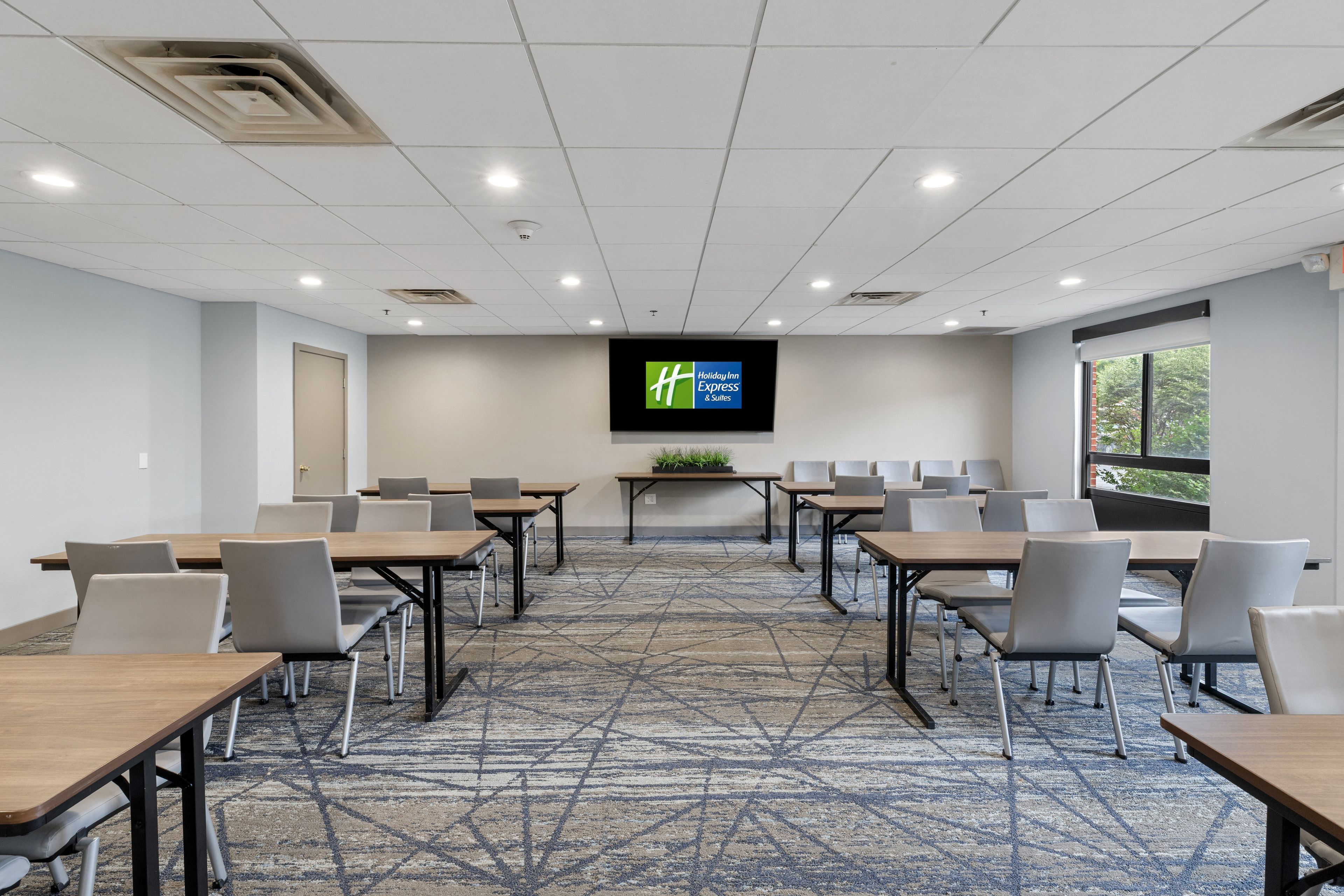 A conference room with a large screen displaying the Holiday Inn Express & Suites logo, multiple tables with chairs, and a patterned carpet.