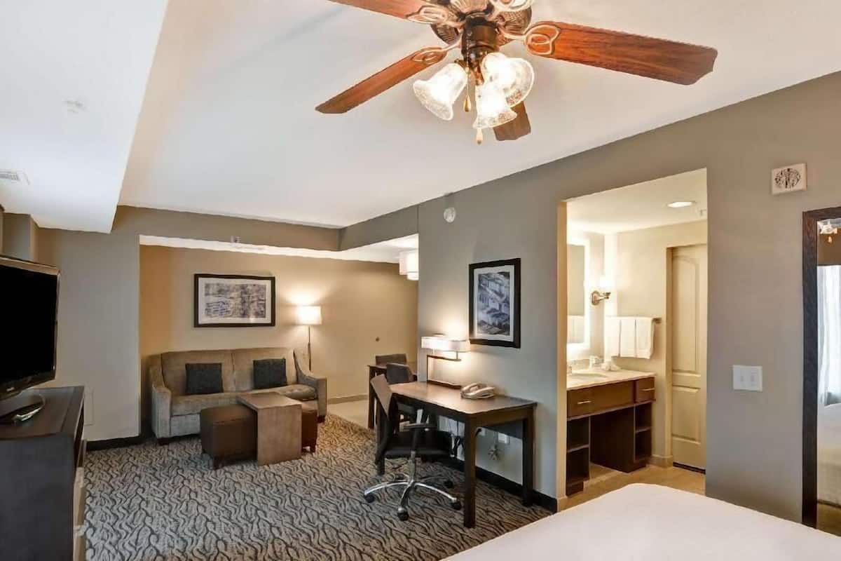 A hotel room with a ceiling fan, a desk, a chair, a sofa, a bed, and a bathroom with a mirror and sink.