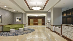 A hotel lobby with a green sofa, two ottomans, a patterned rug, a reception desk, and a large wooden elevator.