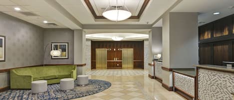 A hotel lobby with a green sofa, two ottomans, a patterned rug, a reception desk, and a large wooden elevator.
