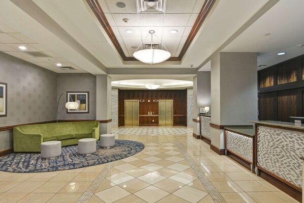 Una hall d'albergo con un divano verde, due poggiapiedi, un tappeto decorato, una reception e un ampio ascensore in legno.
