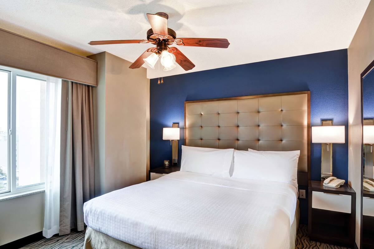 A hotel room with a large bed, a ceiling fan, a nightstand with a lamp, and a window with curtains.