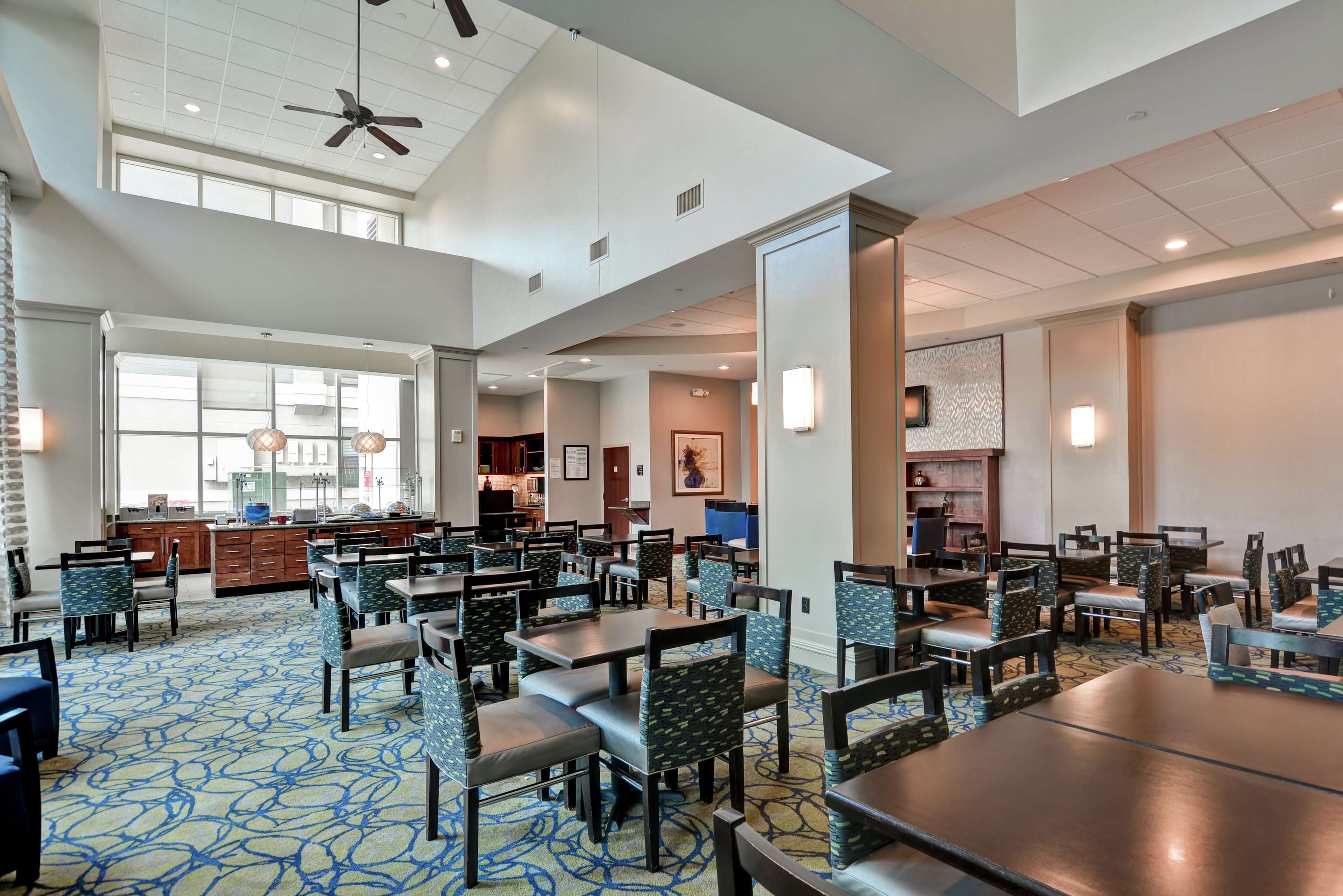 A spacious hotel dining area with multiple tables and chairs, a kitchen in the background, and a high ceiling with a fan.