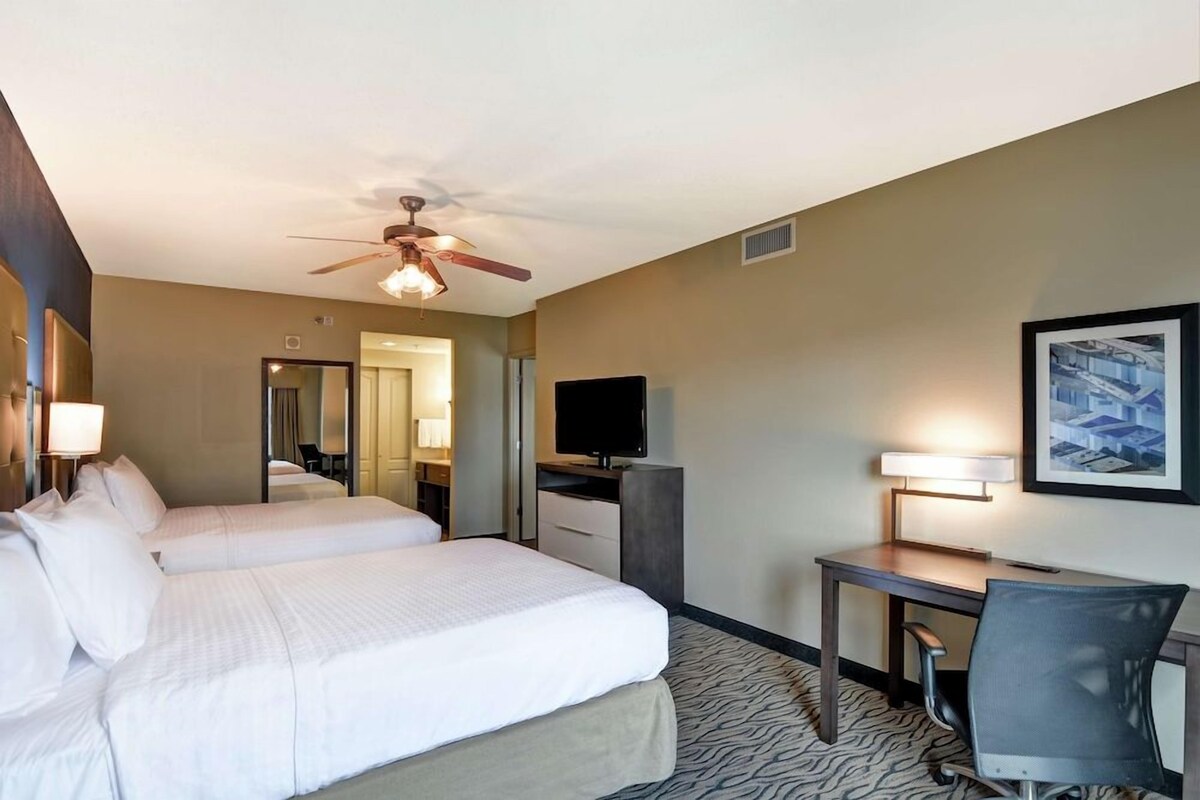 A hotel room with two beds, a desk with a chair, a television, and a ceiling fan.