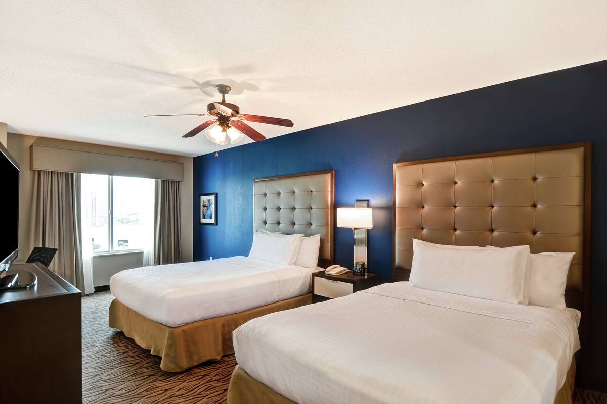 A hotel room with two beds, a ceiling fan, a desk, and a window with curtains.