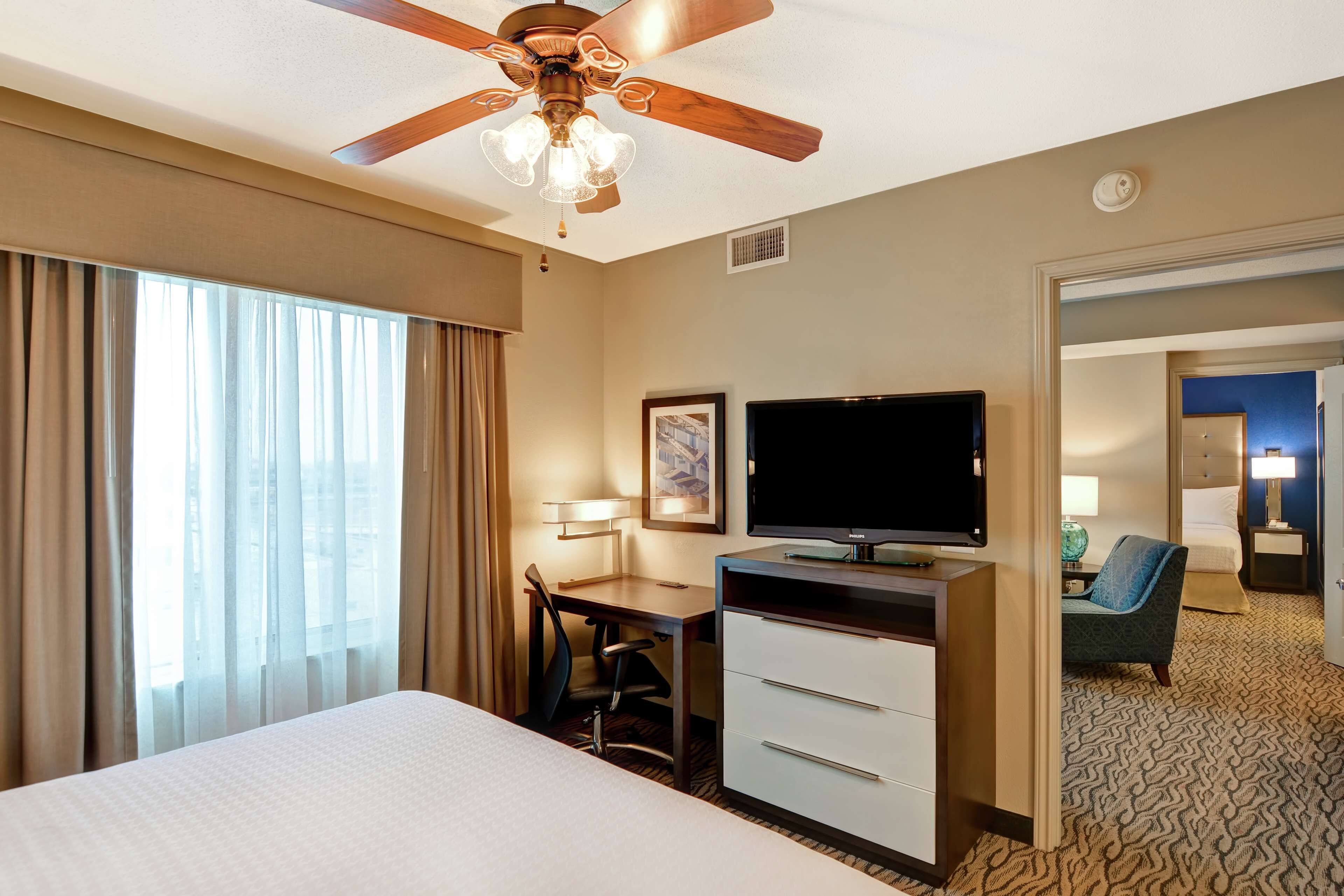 Quarto de hotel com cama, escrivaninha, televisão e ventoinha no teto.