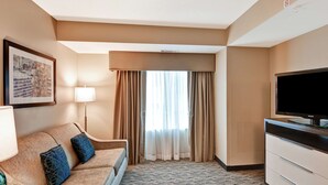 A hotel room with a sofa, a flat-screen TV, a lamp, a framed picture, and a window with curtains.
