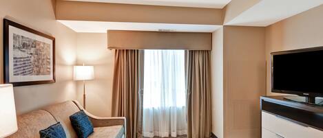 A hotel room with a sofa, a flat-screen TV, a lamp, a framed picture, and a window with curtains.
