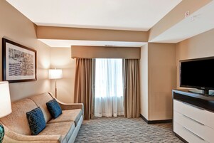 A hotel room with a sofa, a flat-screen TV, a lamp, a framed picture, and a window with curtains.