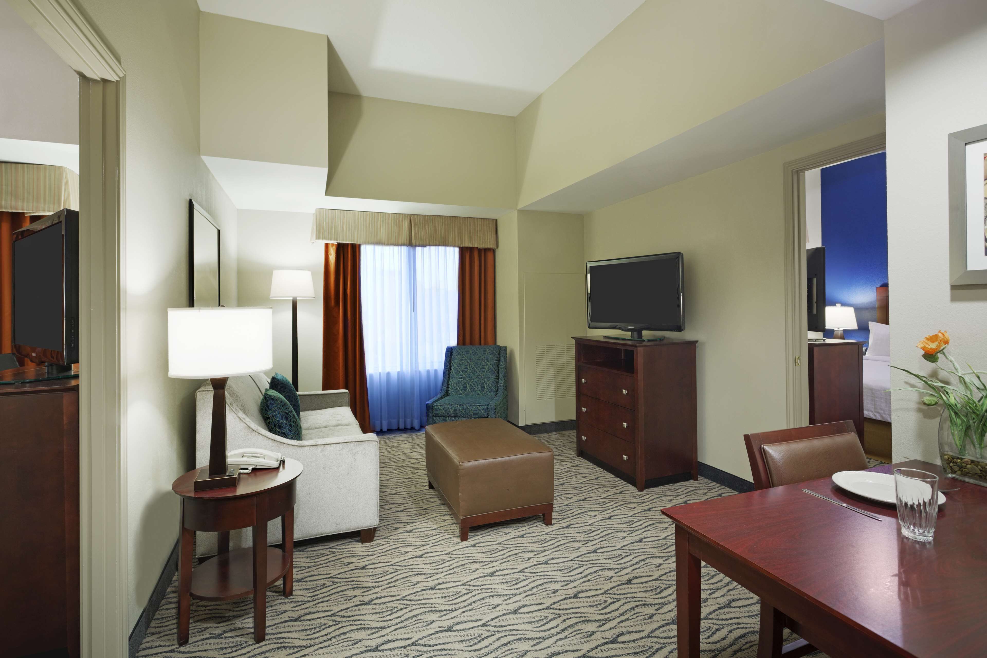 A hotel room with a sofa, armchair, ottoman, TV, and a dining table with a glass of water.