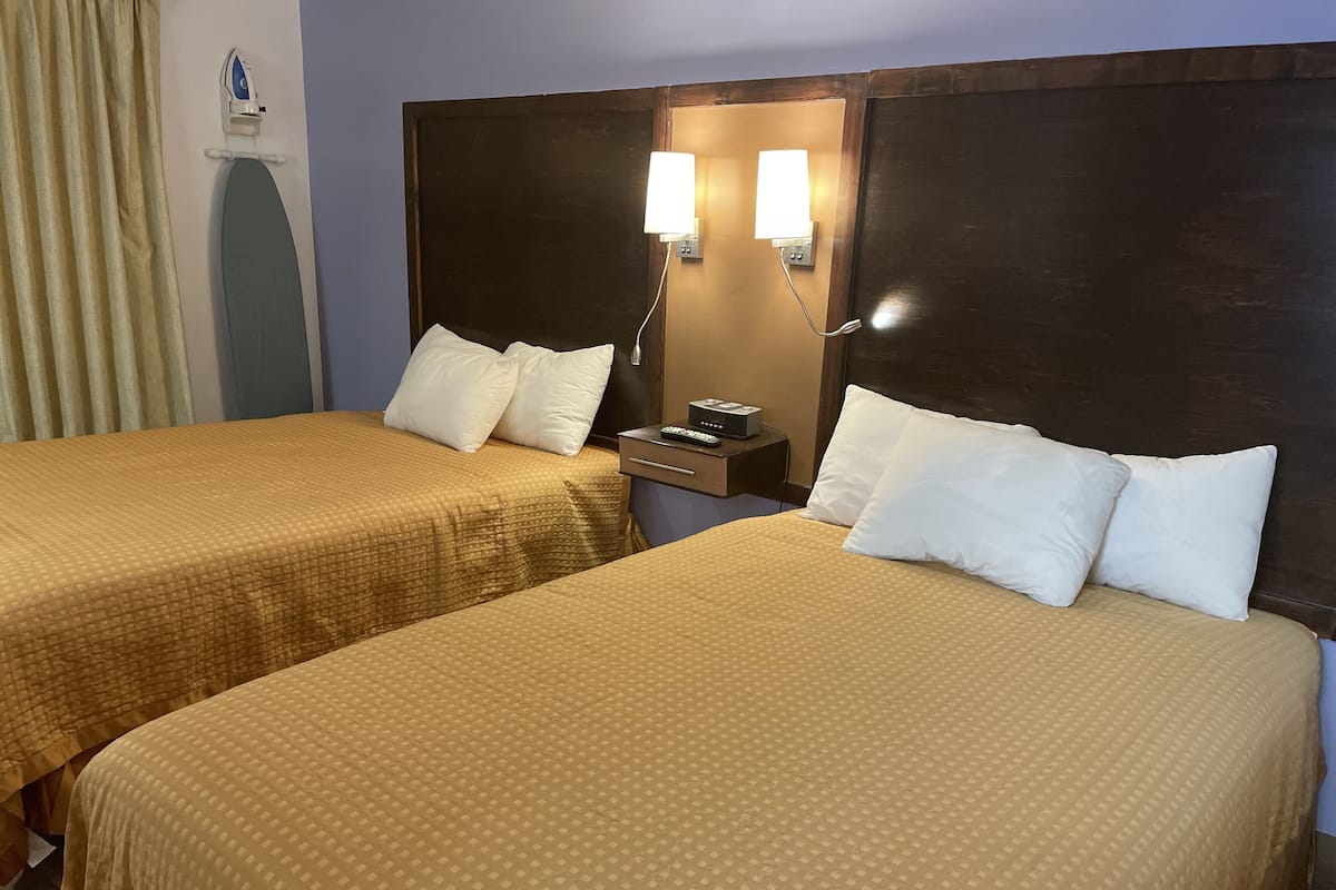 A hotel room with two beds, each with a headboard and pillows. There is a nightstand with a lamp and a remote control.