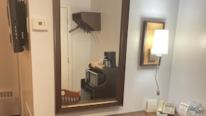 A hotel room with a wooden desk, a chair, a large mirror, a wall-mounted phone, a coffee machine, and a bedside lamp.