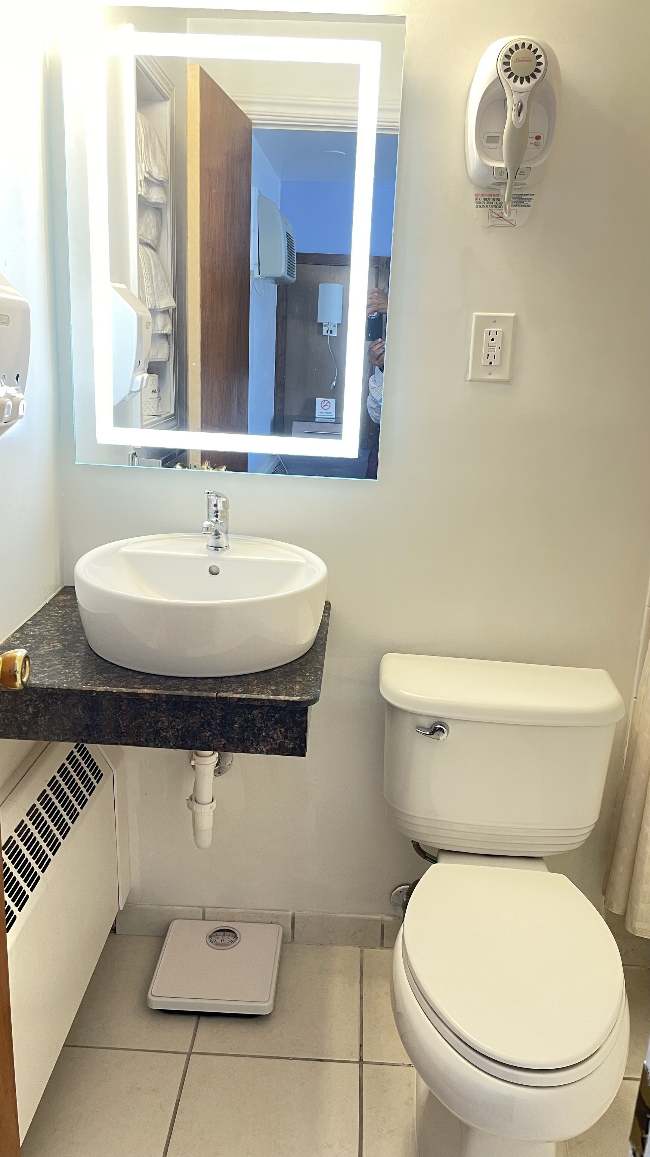 Une salle de bain avec un lavabo blanc, des toilettes, une balance et un sèche-cheveux fixé au mur.