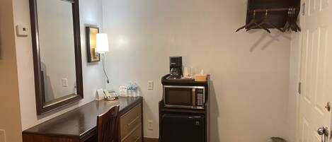 A compact hotel room with a wooden desk, a mirror, a microwave, a small refrigerator, a coffee maker, and a trash bin.