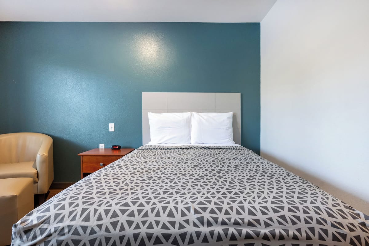 A hotel room with a bed, a chair, a nightstand, and a wall clock.