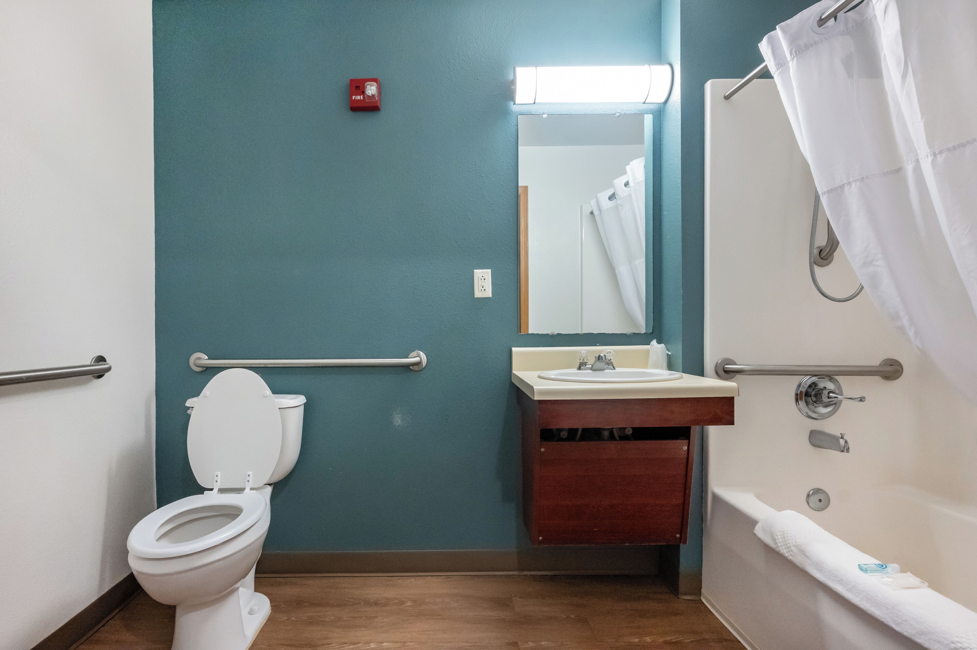 Un baño con inodoro blanco, lavamanos, espejo, ducha con cortina y detector de incendios en la pared.
