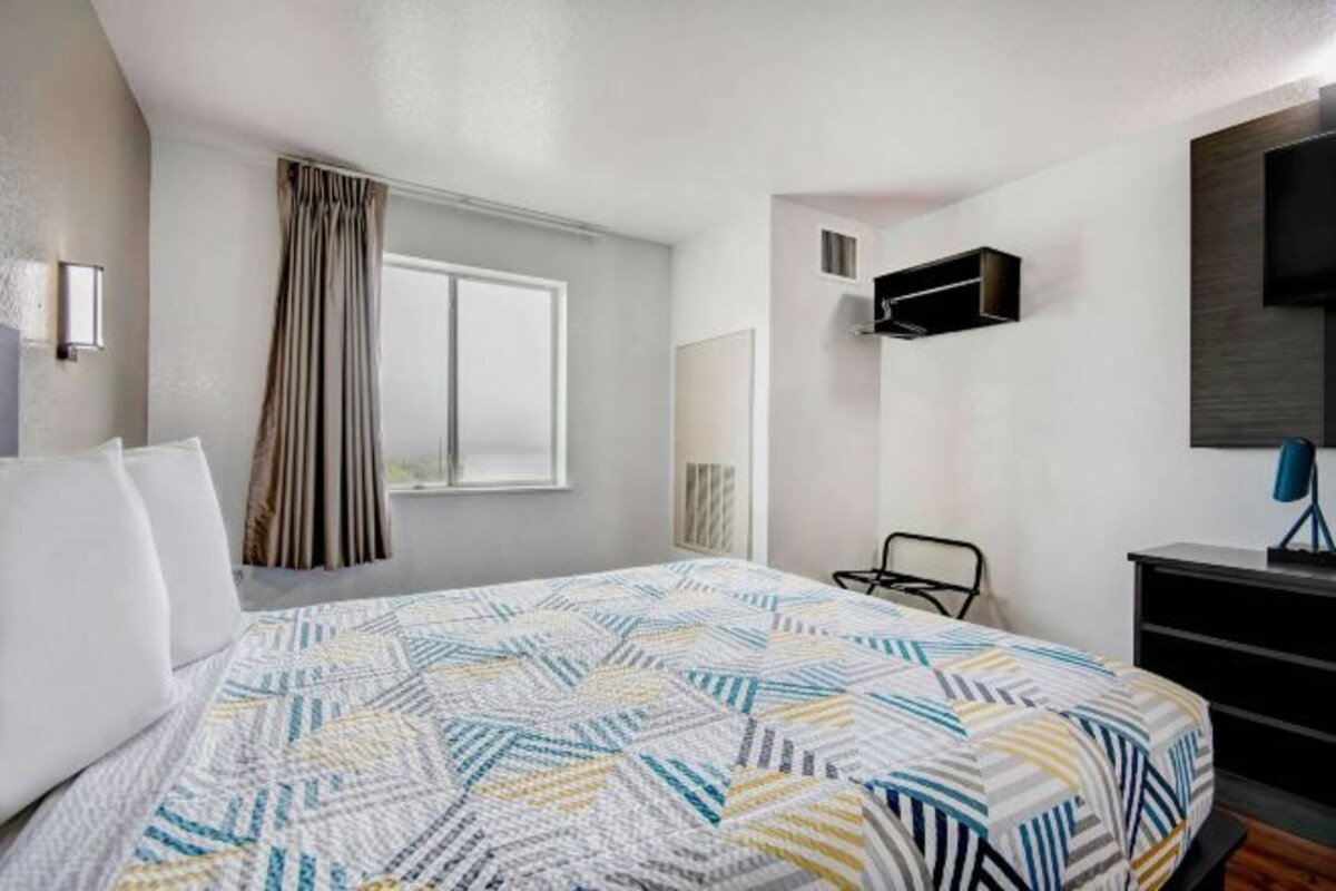 Une chambre d’hôtel équipée d’un lit, d’une fenêtre avec des rideaux, d’un téléviseur à écran plat et d’un climatiseur fixé au mur.