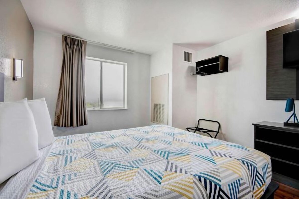 A hotel room with a bed, a TV mounted on the wall, a desk, and a window with curtains.