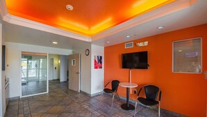 Eine Hotelhalle mit orangefarbenen Wänden, einem gefliesten Boden, einem Flachbildfernseher, zwei Stühlen, einer Uhr und einer Tür zu einem weiteren Raum.