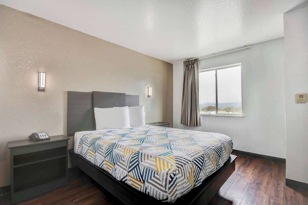 Una habitación de hotel con cama, mesita de noche y ventana con cortinas.