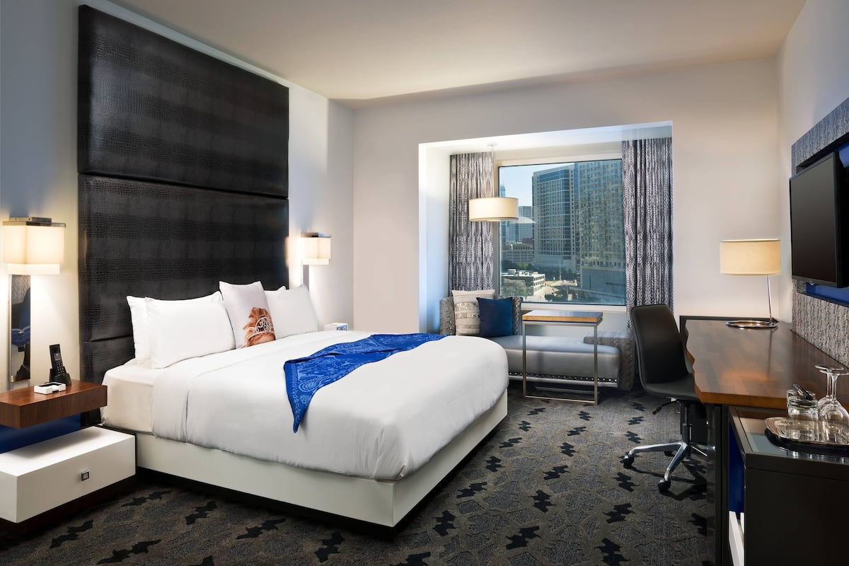 Ein Hotelzimmer mit einem großen Bett, einem Schreibtisch, einem Sessel, einem Fernseher und einem Fenster mit Blick auf die Stadt.