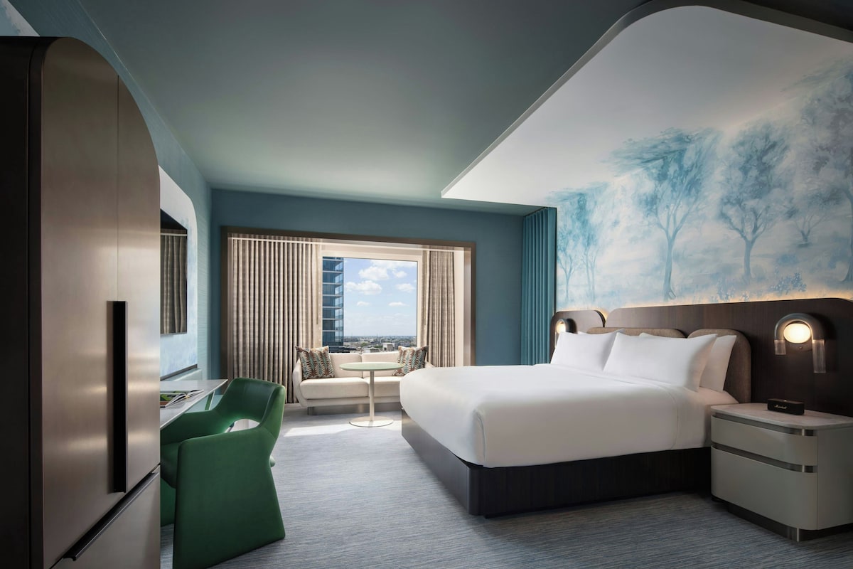 Una camera d'albergo con un letto, una scrivania, una sedia, vista sulla città e un quadro appeso al muro.