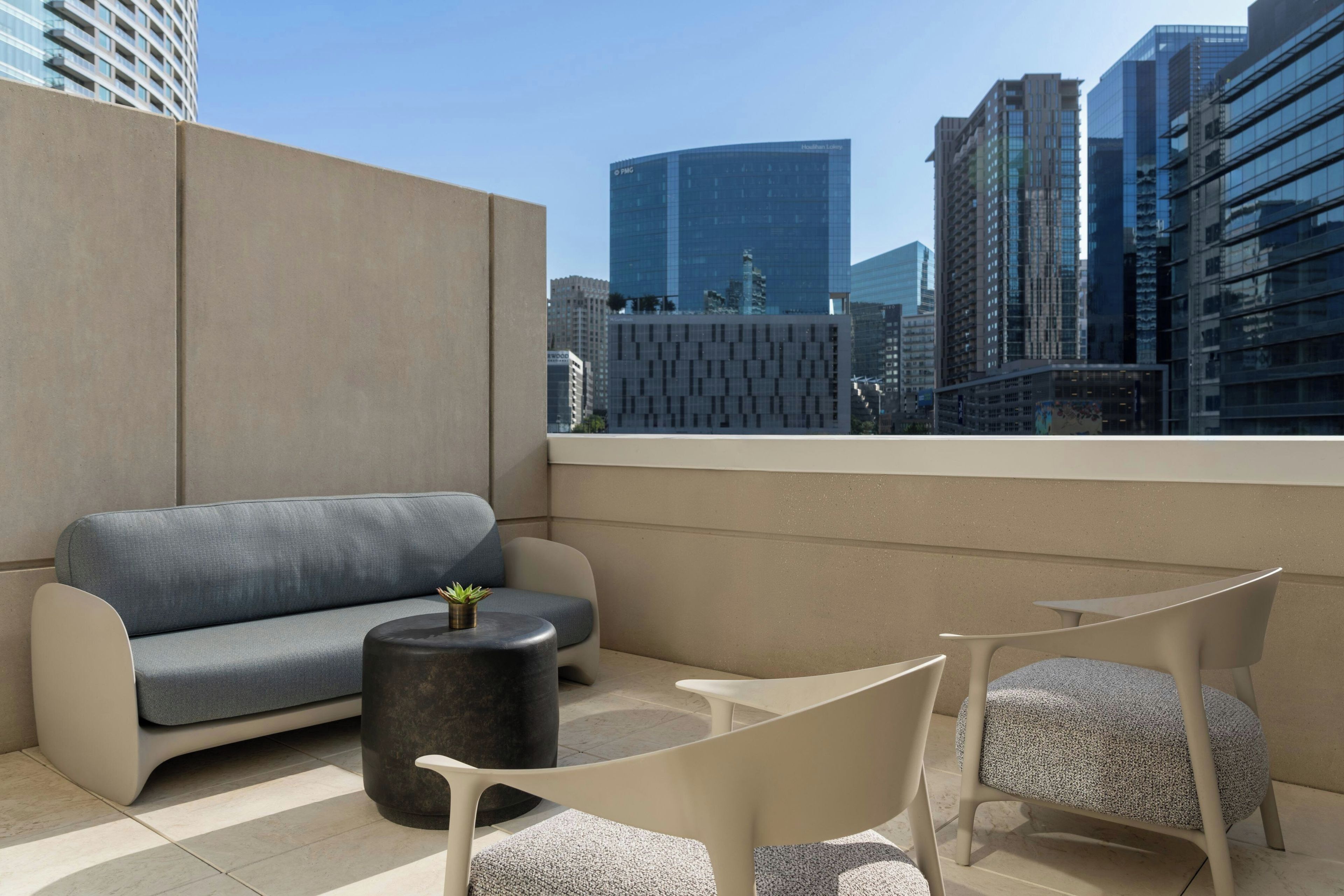 Un área de estar moderna al aire libre con sofá, sillones y una mesita, con vistas a un paisaje urbano con edificios altos.