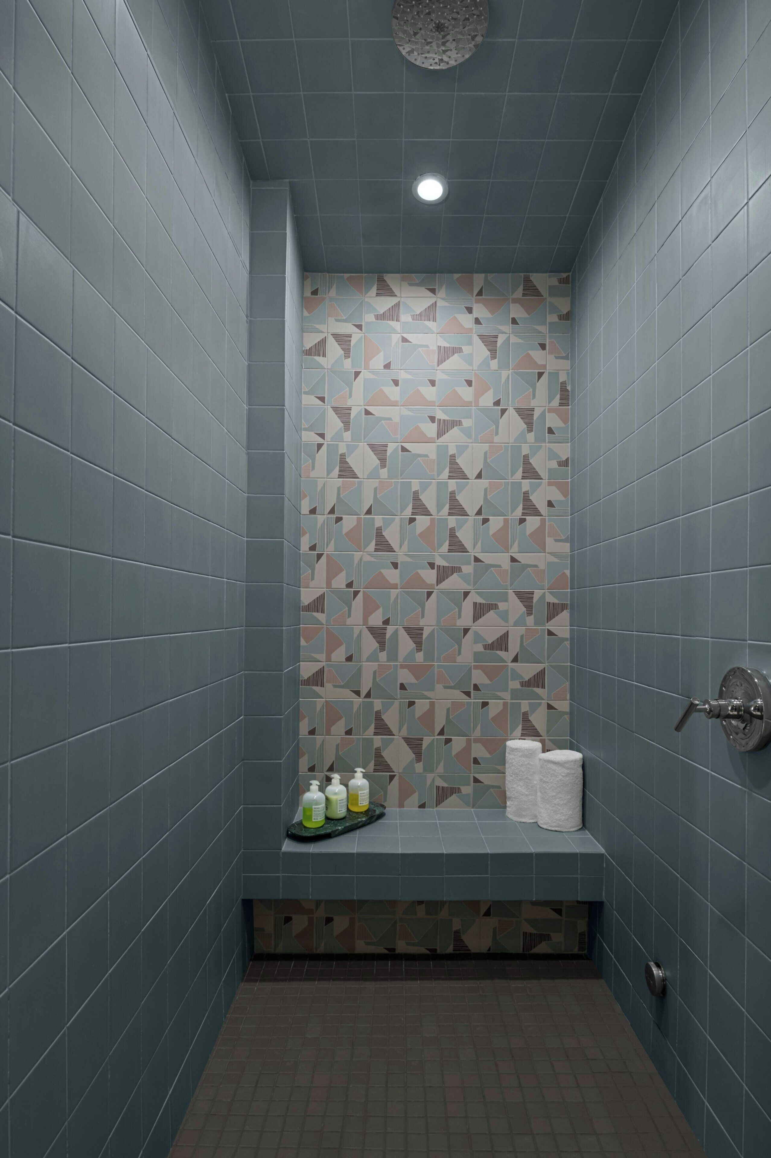 Un espacio de ducha estrecho con azulejos azules, un pared con diseño geométrico y un banco con artículos de aseo.