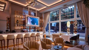 Uma área de bar moderna com uma televisão grande, assentos confortáveis e vista para a rua através de amplas janelas.