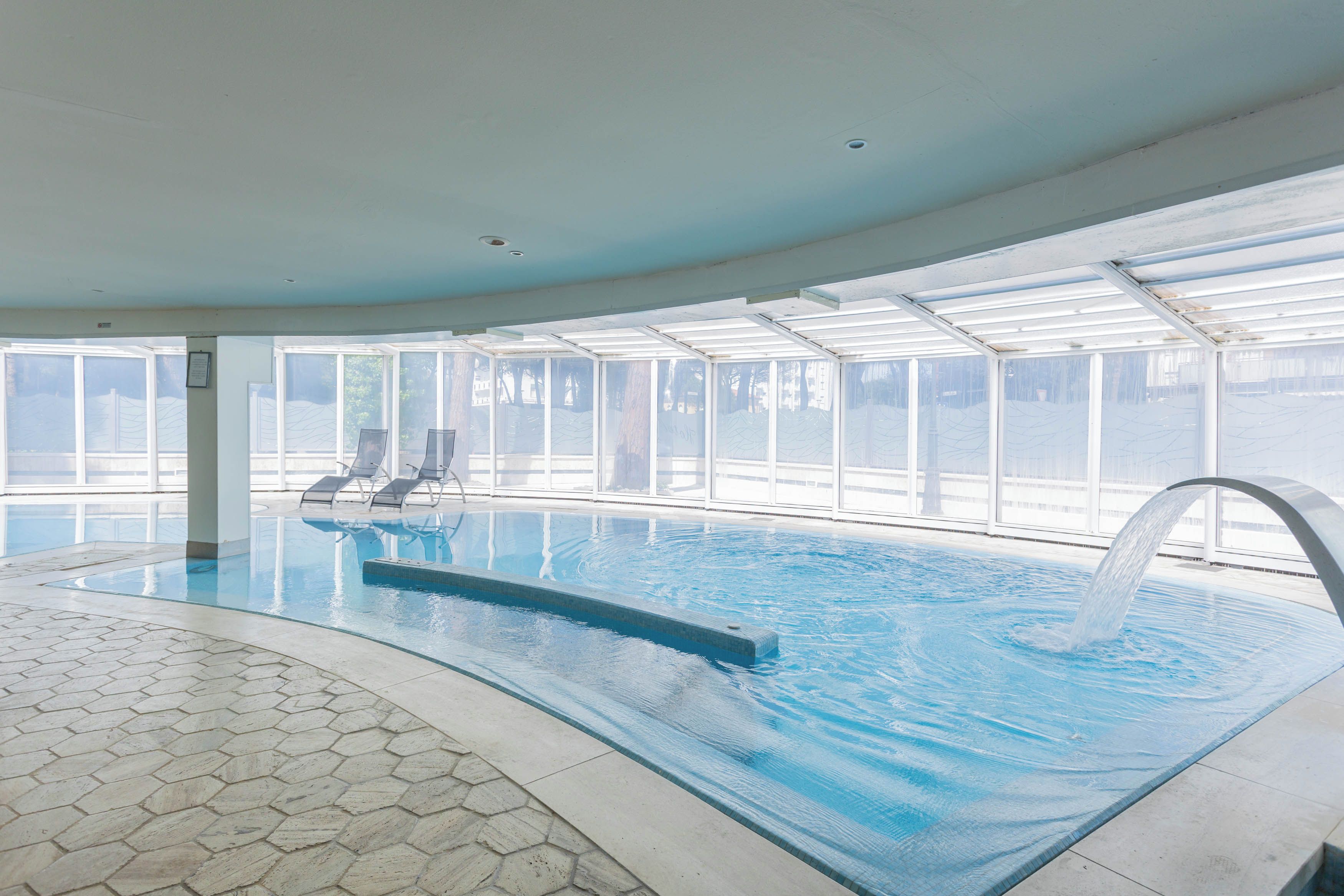 Una piscina interior con paredes de vidrio transparente, suelo de baldosas y una fuente de agua.