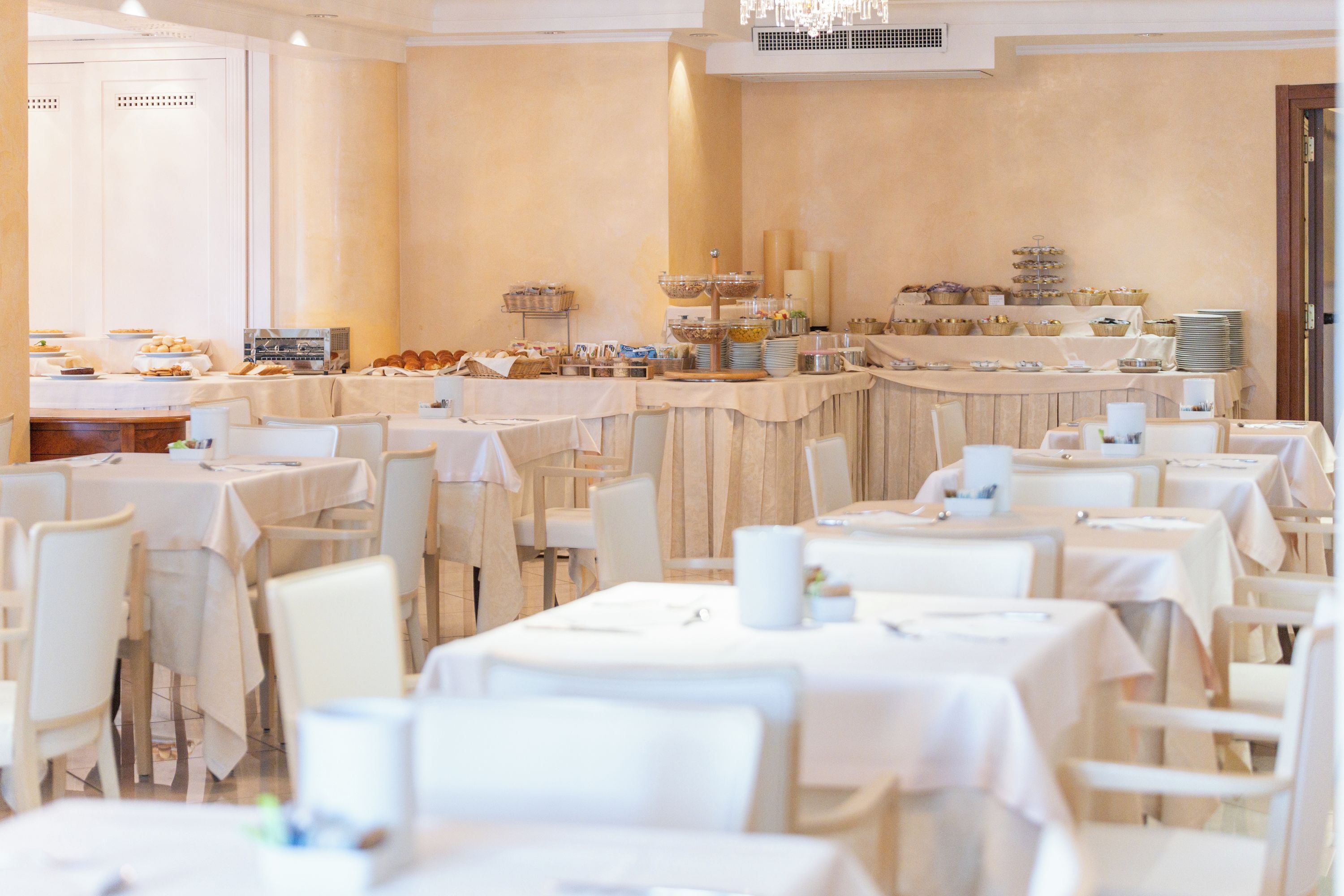 Area ristorazione di un hotel con diversi tavoli e sedie, postazioni per il buffet con varie pietanze e un lampadario.