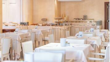 Area ristorazione di un hotel con diversi tavoli e sedie, postazioni per il buffet con varie pietanze e un lampadario.