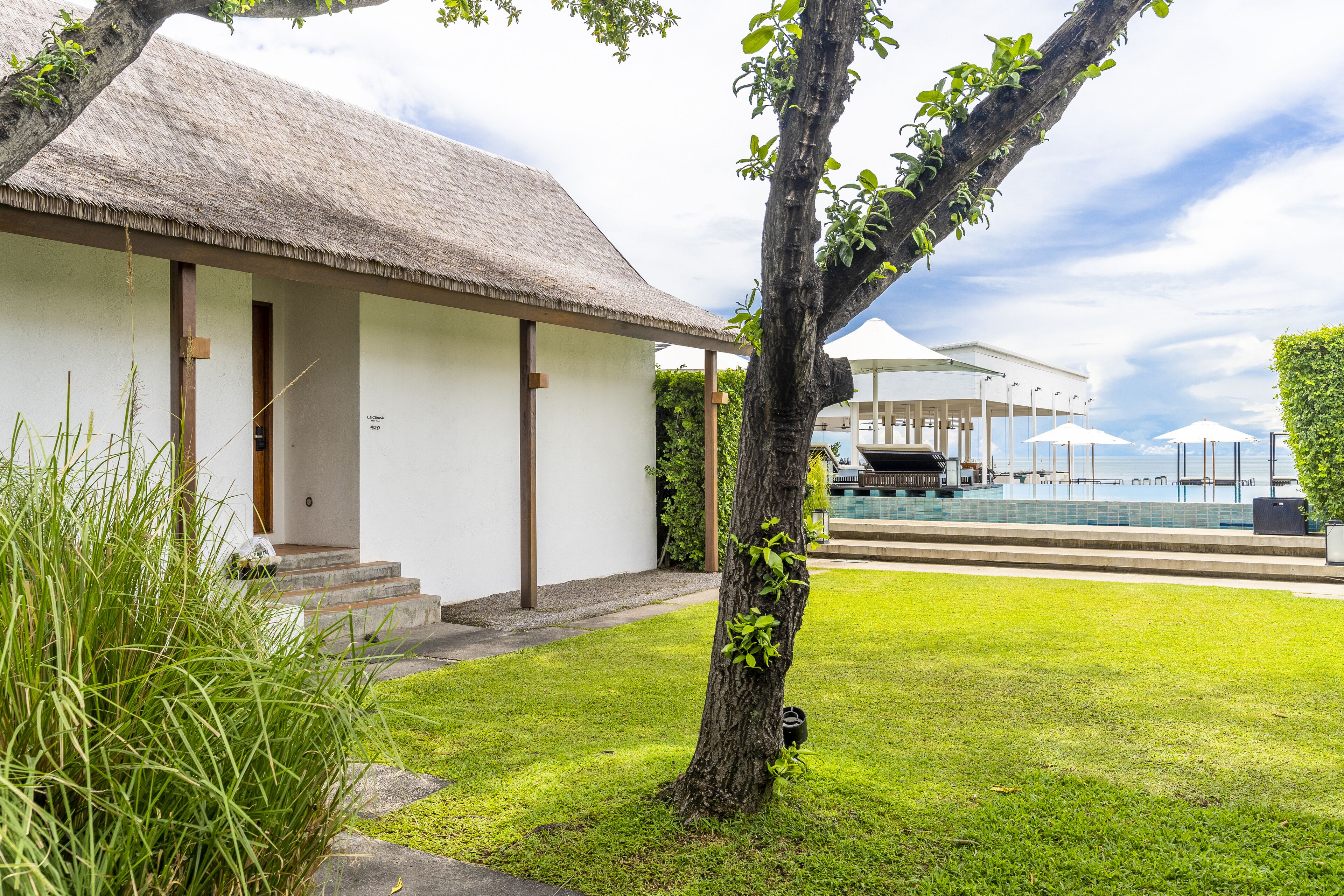 A white building with a thatched roof, a swimming pool, and a covered outdoor area with umbrellas.