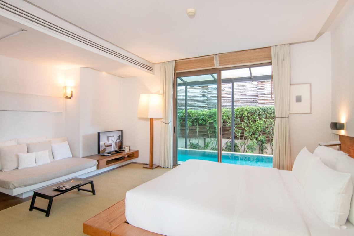A hotel room with a large bed, a sofa, a small table, a TV, and a view of a swimming pool.