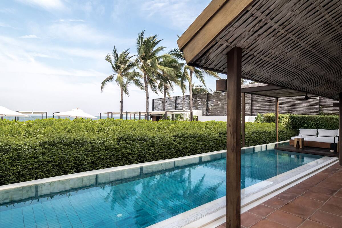 A swimming pool with a tiled edge, surrounded by greenery and palm trees, under a wooden pergola.