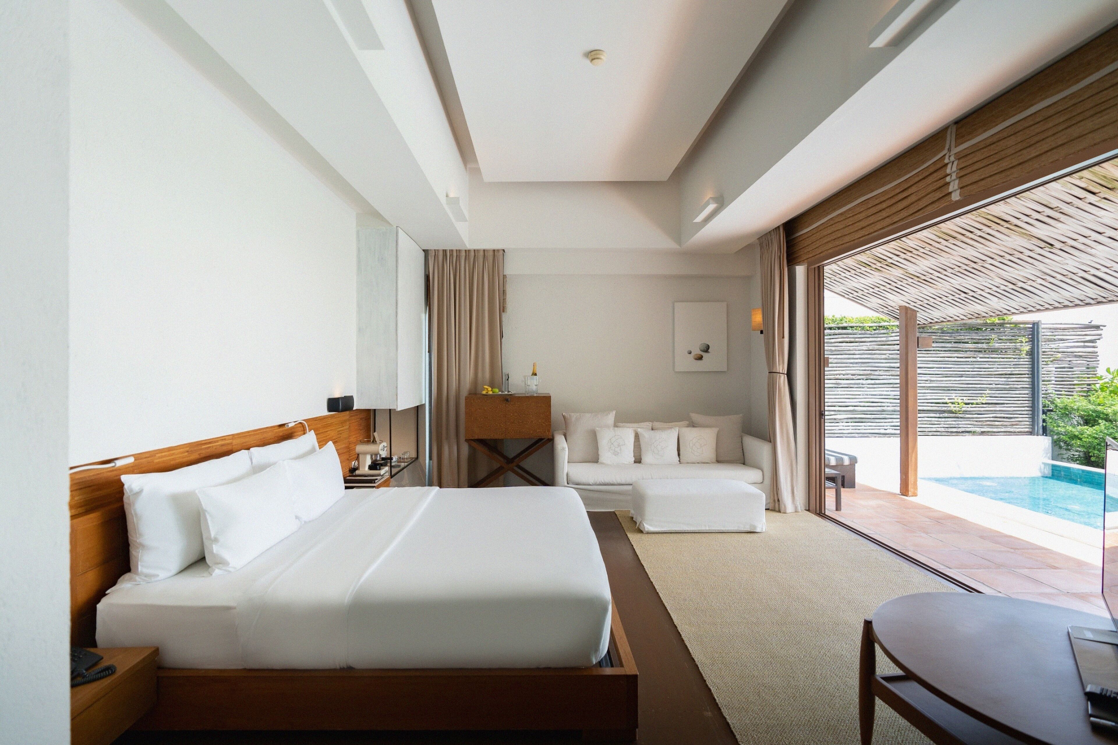 A modern hotel room with a large bed, a sofa, a small table, and a view of an outdoor pool.