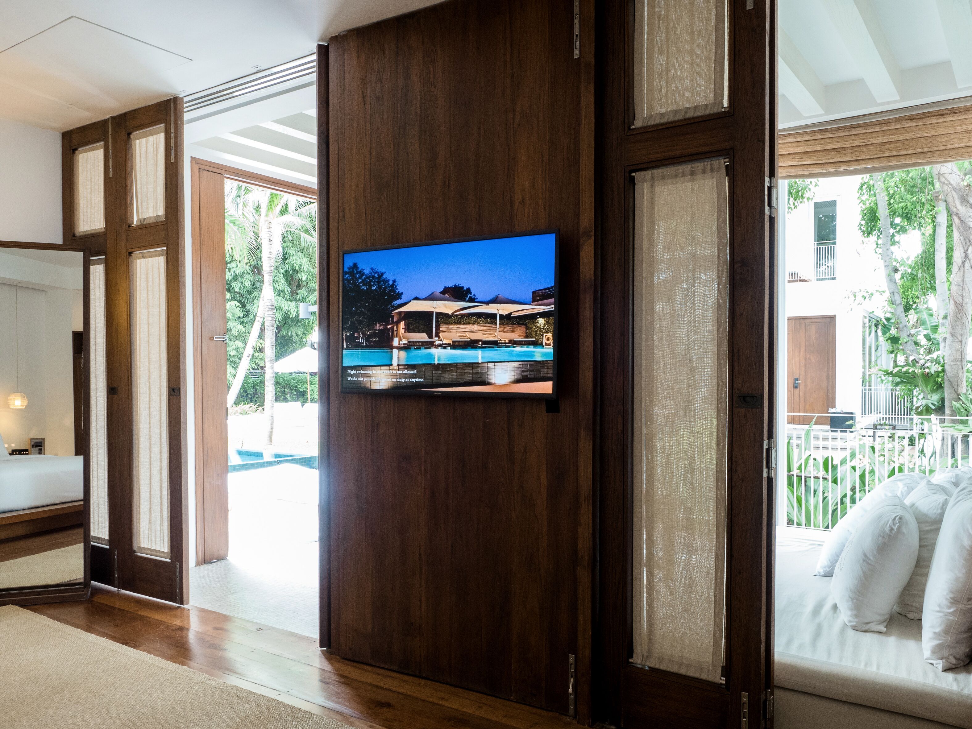 A modern hotel room with a large TV mounted on a wooden panel, a bed with white linens, and a view of a pool and outdoor area.