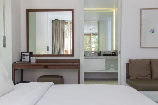 Habitación de hotel con cama, escritorio con computadora, silla, sofá, espejo grande y baño a la vista a través de una puerta abierta.