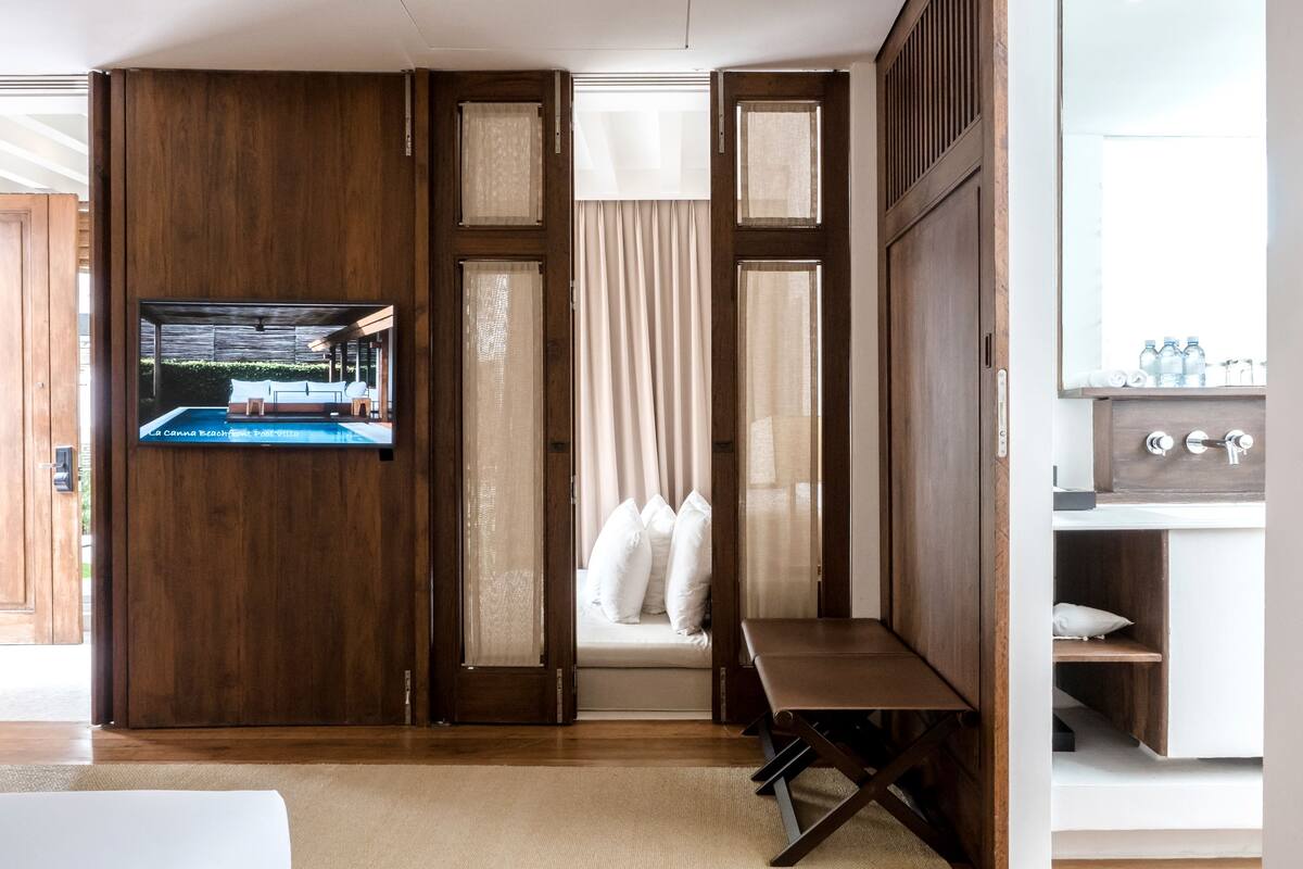 A room with a wooden partition, a television displaying a pool scene, a bed, a bench, and a bathroom with a mirror and sink.