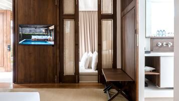 Una habitación con un separador de madera, un televisor mostrando una escena de piscina, una cama, un banco y un baño con espejo y lavamanos.