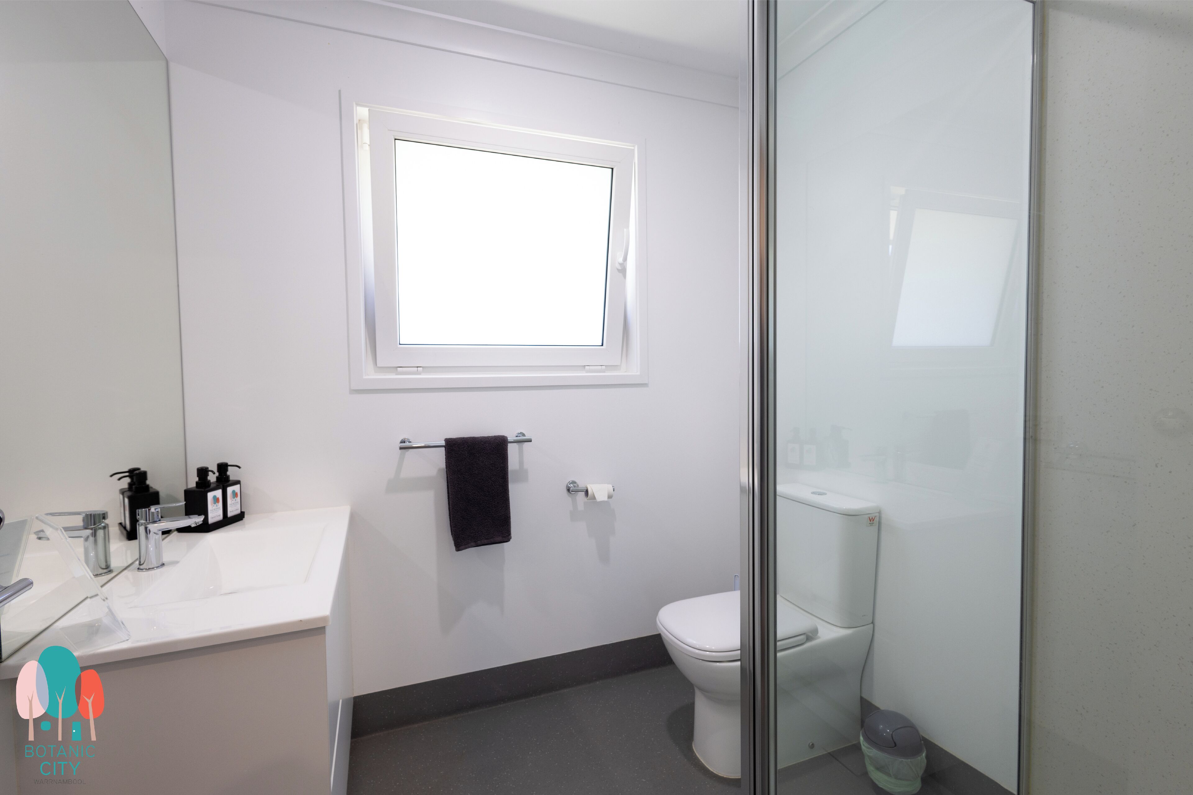 A modern bathroom with a white sink, a toilet, a glass shower enclosure, and a window above the sink.