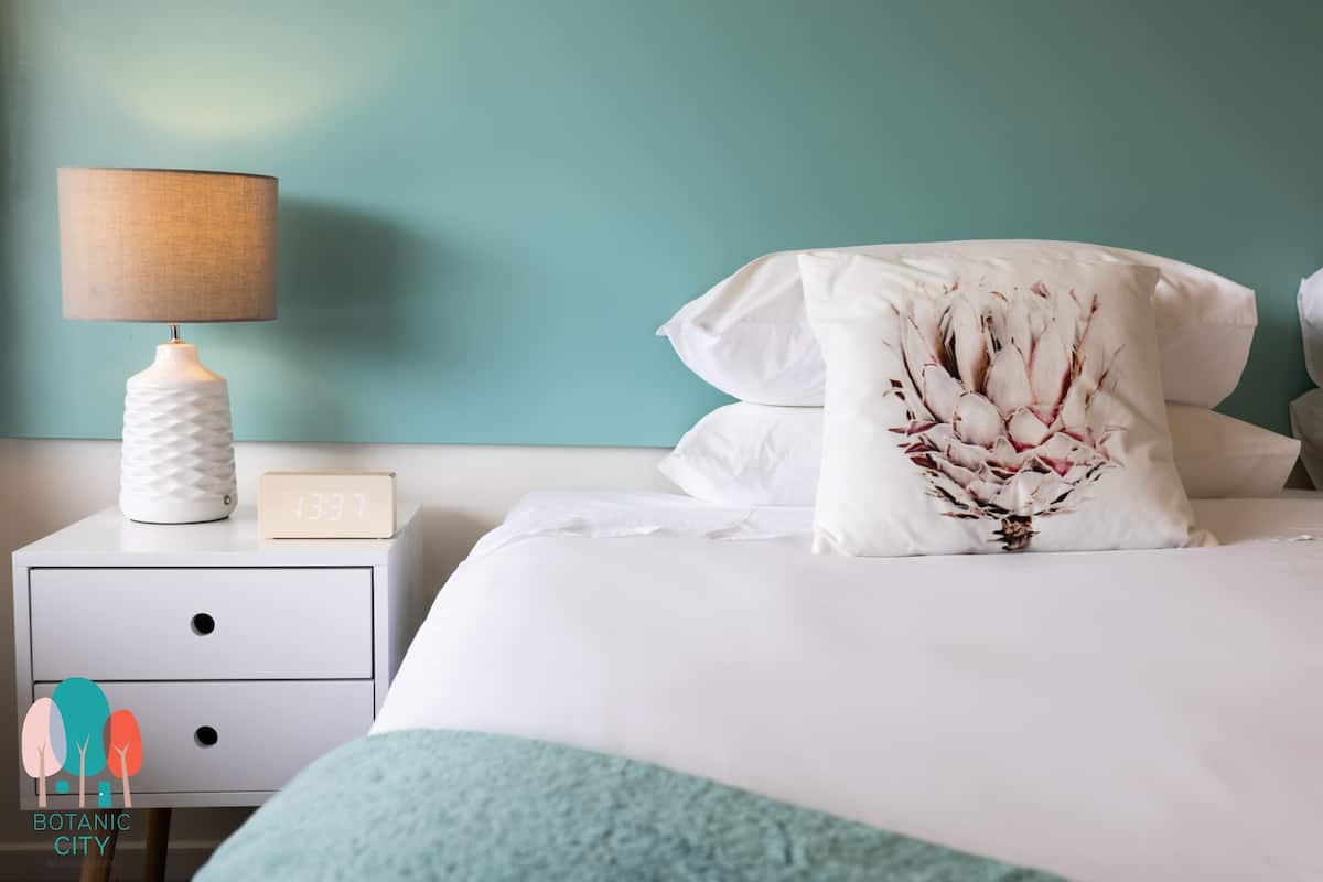 A neatly made bed with white bedding and a decorative pillow, a bedside table with a lamp and a clock, and a teal wall in the background.
