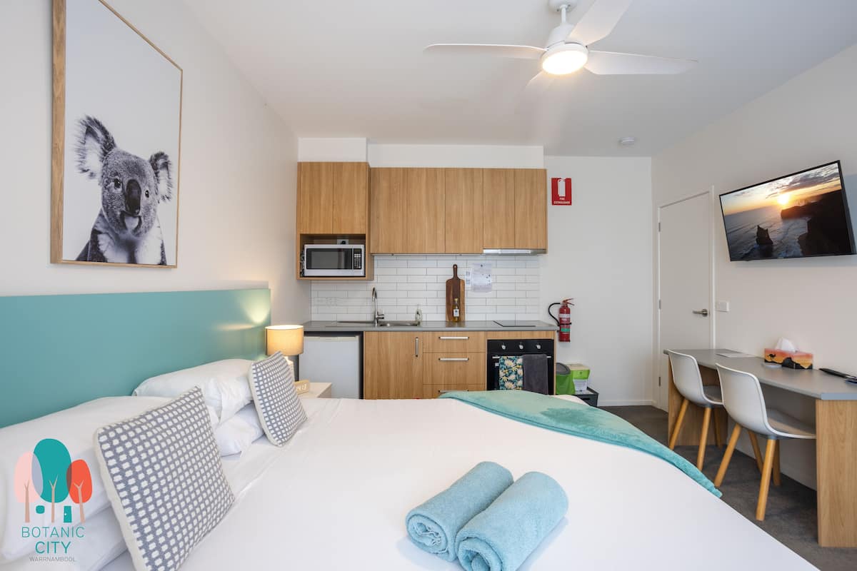 A modern hotel room with a bed, a desk, a kitchenette, and a TV.
