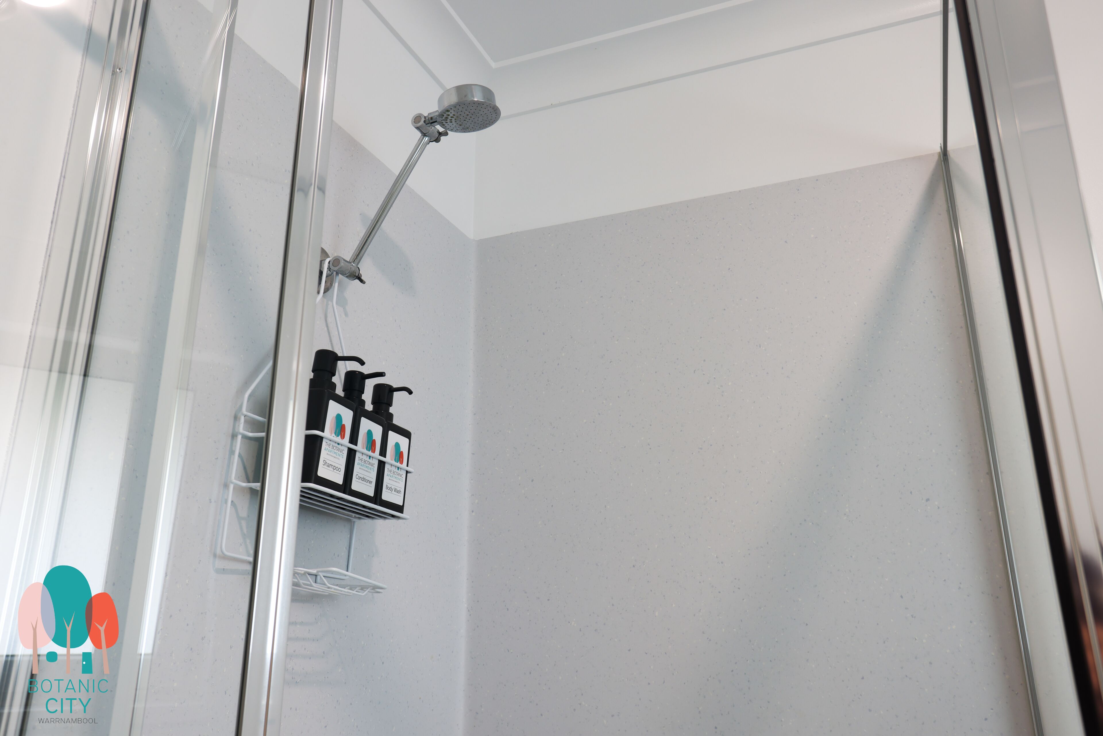 Un espace douche fermé par des parois de verre, doté d’un pommeau de douche à main et d’un distributeur de savon fixé au mur.
