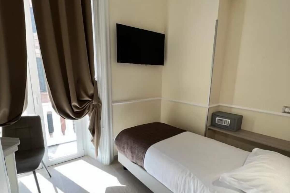 Una habitación de hotel con cama, televisor montado en la pared, mesita de noche con radio y una silla.